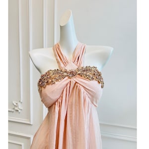 May include: A light peach-colored halter dress with a decorative bodice. The bodice features an ornate design with gold and crystal embellishments. The dress is displayed on a white mannequin against a white wall.