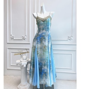 Puede incluir: Un vestido largo con estampado floral azul y blanco, con escote adornado con cuentas y tirantes finos. El vestido tiene una falda fluida con abertura y se exhibe en un maniquí.