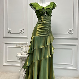 May include: A green, floor-length dress with a ruffled skirt and bodice. The dress features a draped design and a decorative embellishment at the waist. The dress is displayed on a mannequin against a white background.