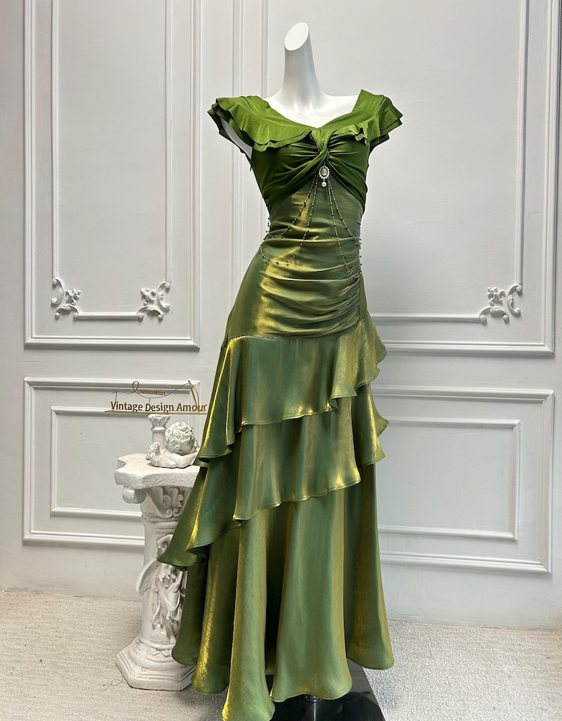 May include: A green, floor-length dress with a ruffled skirt and bodice. The dress features a draped design and a decorative embellishment at the waist. The dress is displayed on a mannequin against a white background.