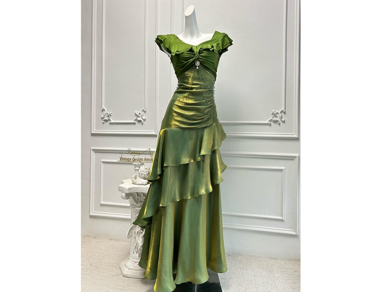 May include: A green satin evening gown with a ruffled skirt and bodice. The dress features a jewel accent at the neckline and is displayed on a mannequin. The text "Vintage Design Amos" is visible.