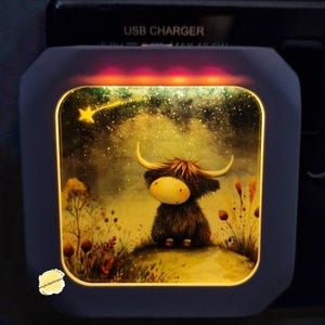 May include: A decorative night light with a Highland cow illustration set against a starry backdrop. The night light has a dark blue frame and emits a warm, yellow light. The text "USB CHARGER" is visible on the top.