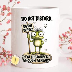 May include: White ceramic mug with a cartoon frog holding a "Do Not Disturb" sign. The frog is green with large eyes and spiky hair. The mug has the text "Do Not Disturb... I'm Disturbed Enough Already."