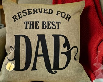 May include: Tan throw pillow with the text "RESERVED FOR THE BEST DAD" in black. A small, round tag is attached to the pillow. A red blanket is draped over the back of the chair. A remote control and a white mug are in the foreground.