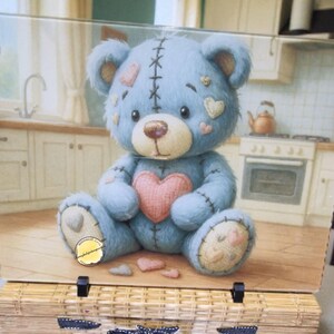 May include: A decorative glass panel features a blue teddy bear with a pink heart and stitched details. The panel is set on a woven basket, creating a charming display. The background shows a kitchen scene, adding a homely touch.