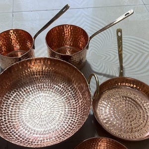 May include: A collection of hammered copper cookware including two saucepans with long handles, a large round deep pan with two side handles, a shallow frying pan with a long handle, and a small bowl, all featuring a polished metallic finish.