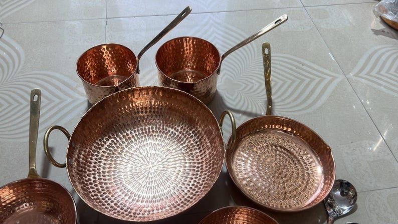 May include: A collection of hammered copper cookware including two saucepans with long handles, a large round deep pan with two side handles, a shallow frying pan with a long handle, and a small bowl, all featuring a polished metallic finish.