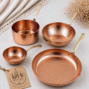 Può includere: Un set di quattro utensili da cucina in rame martellato che include una casseruola profonda, una ciotola arrotondata con manici laterali, un piccolo mestolo e una padella larga, tutti con manici in ottone lucido e un'etichetta DH Crafts.