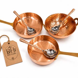 May include: Two copper pans with long brass handles, one round hammered copper bowl with side handles, and three silver-toned spoons. A rectangular brown paper tag with a jute string features the text DH DH Crafts and a small icon.