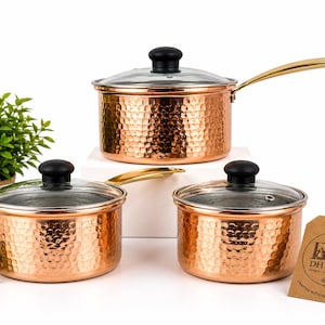 May include: A set of three hammered copper saucepans with gold-toned handles and clear glass lids featuring black knobs. A brown cardboard tag is attached, printed with the text DH Crafts, Crafted with Heritage & Care, and a small logo.