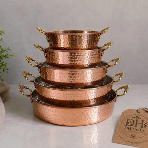 May include: A set of five stacked, round, hammered copper pans with brass handles on each side. The pans decrease in size from bottom to top. A brown paper tag with the text DH Crafts and Crafted with Heritage & Care is attached by a string.