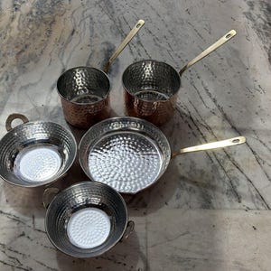 May include: A collection of five hammered metal cookware pieces including two deep saucepans with long handles, one shallow frying pan with a long handle, and two small round bowls with side handles, all featuring a shiny silver interior finish.