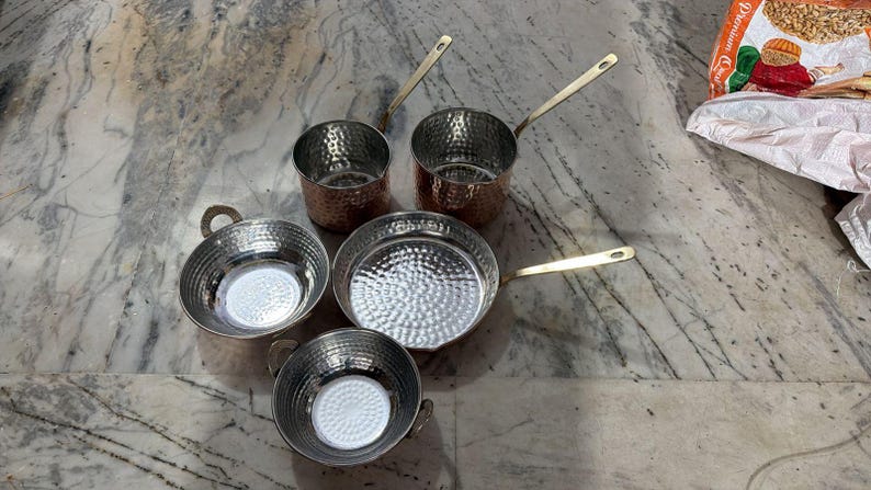 May include: A collection of five hammered metal cookware pieces including two deep saucepans with long handles, one shallow frying pan with a long handle, and two small round bowls with side handles, all featuring a shiny silver interior finish.