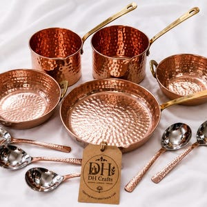 May include: A set of hammered copper cookware including two saucepans with long brass handles, two bowls with side handles, a large frying pan, and five spoons. A brown paper tag is attached, featuring the text DH DH Crafts Crafted with perfection care.