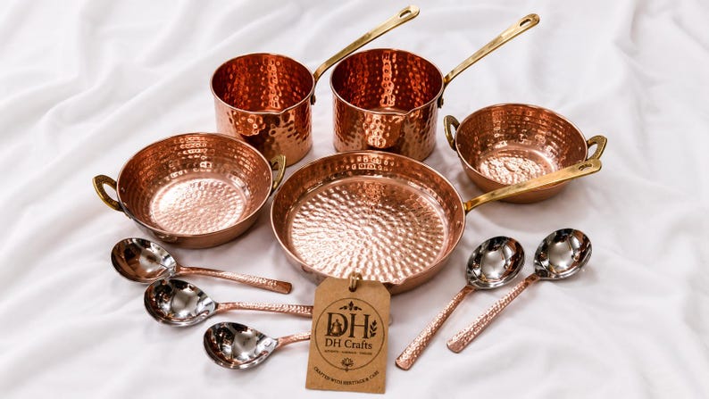 May include: A set of hammered copper cookware including two saucepans with long brass handles, two bowls with side handles, a large frying pan, and five spoons. A brown paper tag is attached, featuring the text DH DH Crafts Crafted with perfection care.