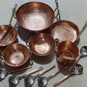 May include: A collection of hammered copper cookware including a large round pot with two handles, three smaller bowls with handles, a shallow pan with a long handle, and a deep cup. Five silver-toned metal spoons with textured handles are included.