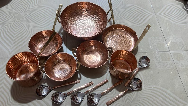 May include: A collection of hammered copper cookware including a large round pot with two handles, three smaller bowls with handles, a shallow pan with a long handle, and a deep cup. Five silver-toned metal spoons with textured handles are included.