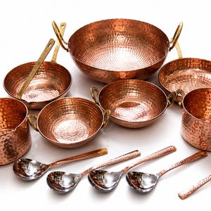 May include: A set of hammered copper cookware including various bowls, pans, and spoons with metallic handles. A brown cardboard tag is present with the text DH Crafts and Crafted with Heritage & Care. The items feature a textured, dimpled surface.