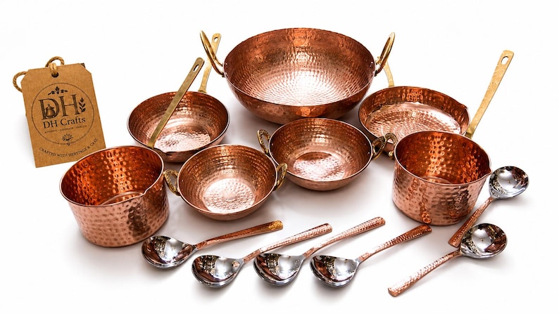 May include: A set of hammered copper cookware including various bowls, pans, and spoons with metallic handles. A brown cardboard tag is present with the text DH Crafts and Crafted with Heritage & Care. The items feature a textured, dimpled surface.