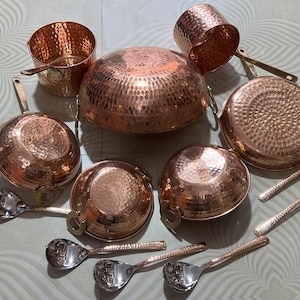 May include: A collection of hammered copper cookware including a large central bowl, two handled pans, two small bowls, and a cylindrical container, accompanied by five silver-toned metal spoons with long handles arranged in a circular formation.