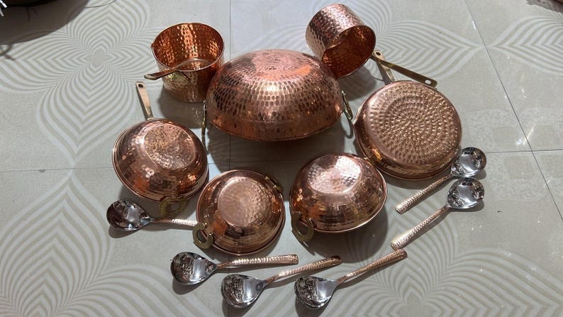 May include: A collection of hammered copper cookware including a large central bowl, two handled pans, two small bowls, and a cylindrical container, accompanied by five silver-toned metal spoons with long handles arranged in a circular formation.