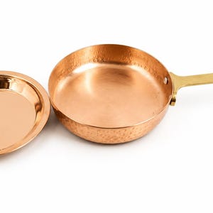 May include: A round copper frying pan with a hammered exterior finish and a long, flat brass handle. The pan is accompanied by a matching circular copper lid that features a small, centred brass knob for lifting and handling the cover.