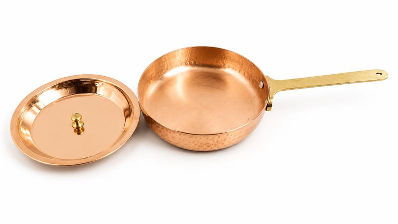 May include: A round copper frying pan with a hammered exterior finish and a long, flat brass handle. The pan is accompanied by a matching circular copper lid that features a small, centred brass knob for lifting and handling the cover.