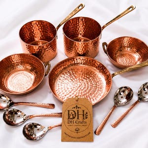 May include: A set of hammered copper cookware including two saucepans with long handles, two bowls with side handles, one frying pan, and four spoons with metallic bowls. A brown rectangular tag features the text DH Crafts and DH logo.