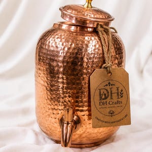 May include: A hammered copper water dispenser with a matching lid, a gold-toned knob, and a copper spigot. A brown paper tag is tied to the neck with twine, featuring the text DH Crafts, Authentic Handmade Timeless, and Crafted with Heritage & Care.