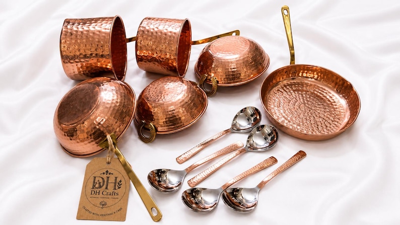 May include: A set of hammered copper kitchenware including two mugs, three bowls with brass handles, a frying pan with a long brass handle, and four spoons with copper-toned handles and silver-colored bowls. A brown tag displays the text DH Crafts.