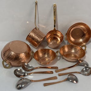 May include: A collection of hammered copper cookware including a large bowl, two smaller bowls with side handles, two saucepans with long brass handles, and six silver-toned metal spoons with copper-colored handles arranged on a white surface.