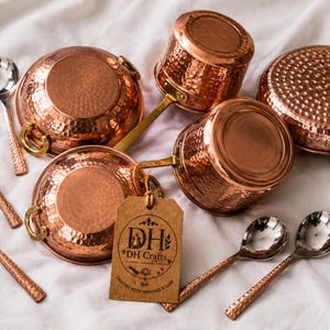 May include: A collection of hammered copper cookware including two round pots with side handles, two saucepans with long handles, and five silver-toned spoons with textured copper handles. A brown paper tag displays the text DH DH Crafts.