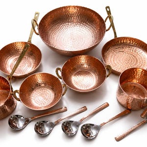 May include: A set of hammered copper cookware including a large bowl, two smaller bowls, two saucepans with long handles, and a deep pot. Six silver-toned metal spoons are arranged below. A brown cardboard tag displays the text DH Crafts.