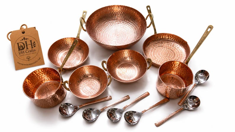 May include: A set of hammered copper cookware including a large bowl, two smaller bowls, two saucepans with long handles, and a deep pot. Six silver-toned metal spoons are arranged below. A brown cardboard tag displays the text DH Crafts.