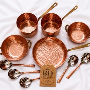 May include: A set of hammered copper cookware including two saucepans with long handles, two bowls with side handles, one frying pan, and five spoons. A brown paper tag with the text DH DH Crafts is attached to the frying pan handle.