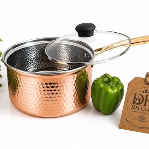 May include: A hammered copper saucepan with a gold-toned handle and a clear glass lid featuring a black knob. Next to the pan sits a green bell pepper and a brown cardboard tag printed with the text DH Crafts, Crafted with Heritage & Care.