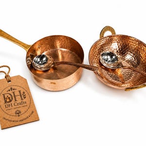 May include: A set of two hammered copper cooking vessels, one with a long handle and one with two side handles, each containing a silver-toned metal spoon. A brown paper tag with the text DH Crafts, Crafted with Heritage & Care is attached.