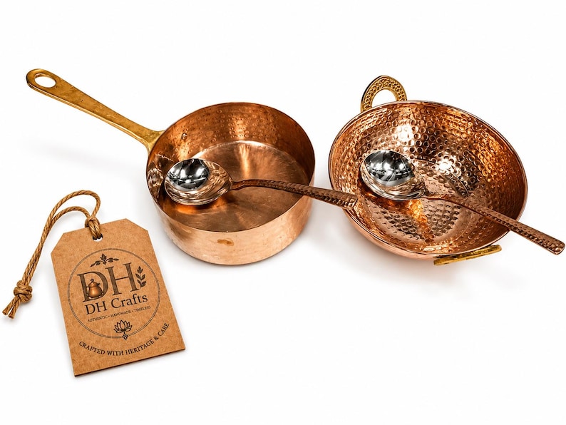 May include: A set of two hammered copper cooking vessels, one with a long handle and one with two side handles, each containing a silver-toned metal spoon. A brown paper tag with the text DH Crafts, Crafted with Heritage & Care is attached.