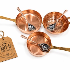 May include: A set of three copper kitchen vessels including two pans with long brass handles and one bowl with side handles, each containing a silver-toned spoon with a textured handle. A brown paper tag displays the text DH Crafts Crafted with Heritage & Care.