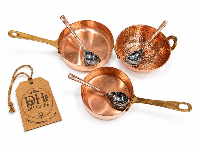 May include: A set of three copper kitchen vessels including two pans with long brass handles and one bowl with side handles, each containing a silver-toned spoon with a textured handle. A brown paper tag displays the text DH Crafts Crafted with Heritage & Care.