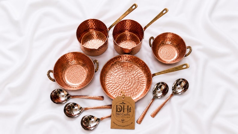 May include: A set of hammered copper cookware including two saucepans with long handles, two bowls with side handles, one frying pan, and five spoons. A brown paper tag with the text DH DH Crafts is attached to the frying pan handle.