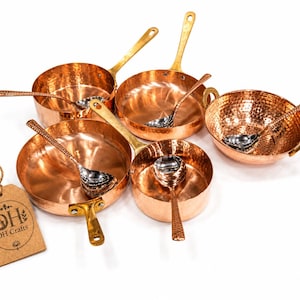 May include: A set of hammered copper cookware including four pans and a bowl, each paired with a matching silver-toned spoon. A brown rectangular paper tag with the text DH DH Crafts is attached by a string to the largest pan in the collection.