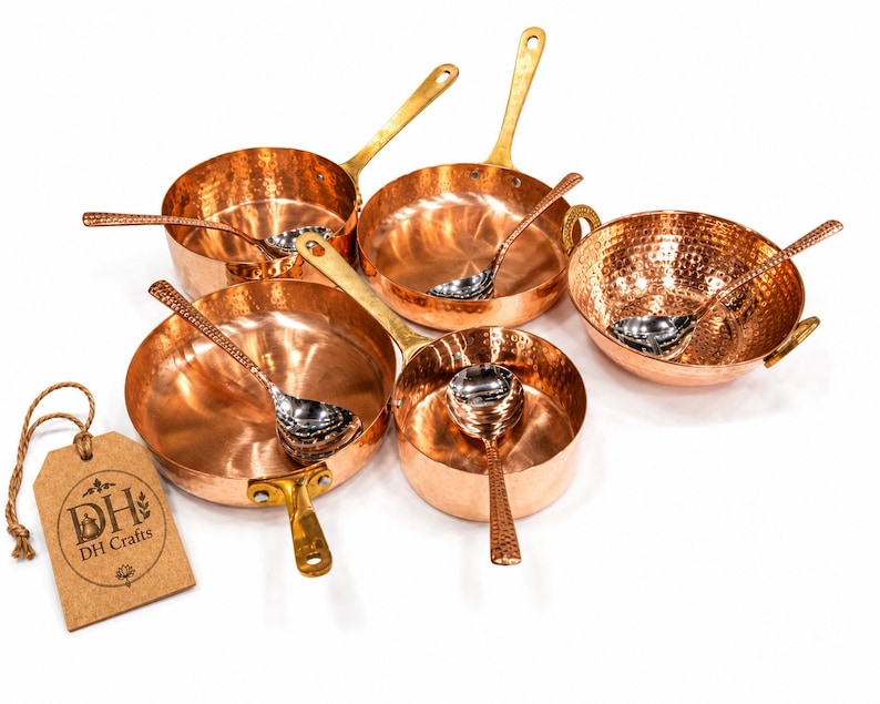 May include: A set of hammered copper cookware including four pans and a bowl, each paired with a matching silver-toned spoon. A brown rectangular paper tag with the text DH DH Crafts is attached by a string to the largest pan in the collection.