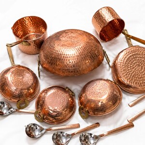 May include: A set of hammered copper cookware including a large central bowl, two saucepans with handles, a small cup, and four silver-toned metal spoons. A brown rectangular tag with the text DH Crafts and Crafted with Heritage & Care is included.