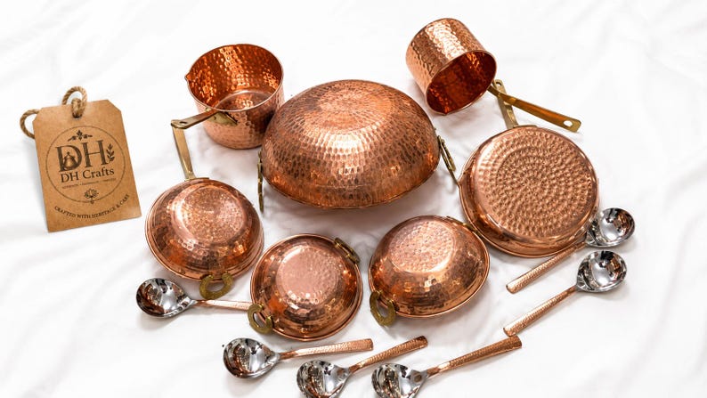 May include: A set of hammered copper cookware including a large central bowl, two saucepans with handles, a small cup, and four silver-toned metal spoons. A brown rectangular tag with the text DH Crafts and Crafted with Heritage & Care is included.