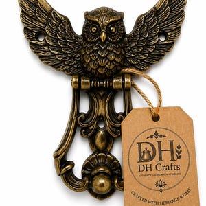 Op de afbeelding: Een bronskleurige metalen deurklopper in de vorm van een uil met gespreide vleugels, voorzien van gedetailleerde veren en een ronde basis. Een bruin papieren label is met touw bevestigd en bevat de tekst DH DH Crafts Authentic Handmade Timeless.