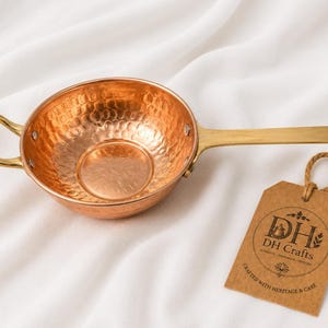 May include: A hammered copper pan with a polished brass handle and a small side loop handle. The pan features a brown paper tag attached with a jute string, displaying the text DH DH Crafts Crafted with Heritage & Care in black ink.