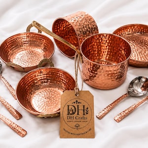 May include: A set of hammered copper kitchenware including two bowls, two pots, and five spoons with metallic handles. A brown cardboard tag is attached to one pot with twine, featuring the printed text DH Crafts and Crafted with Heritage & Care.