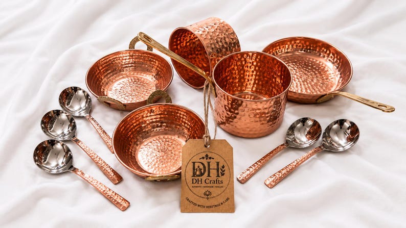 May include: A set of hammered copper kitchenware including two bowls, two pots, and five spoons with metallic handles. A brown cardboard tag is attached to one pot with twine, featuring the printed text DH Crafts and Crafted with Heritage & Care.