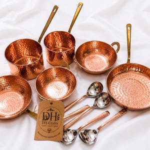 May include: A collection of hammered copper cookware including two saucepans with long handles, two bowls with side handles, one frying pan with a long handle, and four spoons. A brown paper tag with the text DH Crafts is attached to the set.